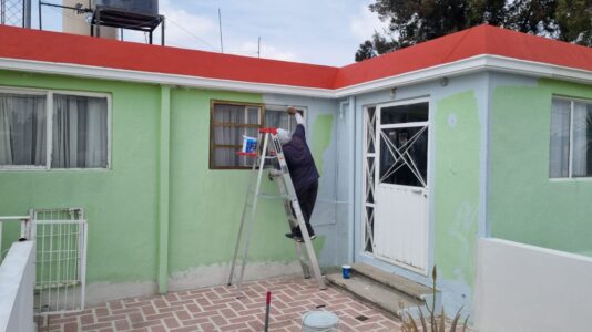 Proceso de pintura exterior por M2 Arquitectos: técnico sobre escalera aplicando pintura azul claro sobre un muro verde para la renovación de una fachada residencial.