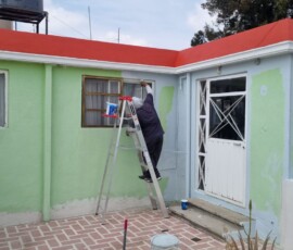 Proceso de pintura exterior por M2 Arquitectos: técnico sobre escalera aplicando pintura azul claro sobre un muro verde para la renovación de una fachada residencial.