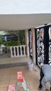 Proceso de remodelación de terraza residencial por M2 Arquitectos, mostrando una escalera de tijera, materiales de construcción y un portón de herrería negra con diseño artístico al fondo.