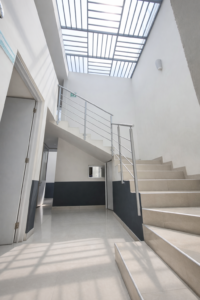 Escalera moderna de concreto con barandal de acero inoxidable en el interior de un edificio iluminado por un tragaluz de rejilla metálica.