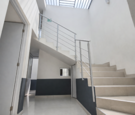 Escalera moderna de concreto con barandal de acero inoxidable en el interior de un edificio iluminado por un tragaluz de rejilla metálica.