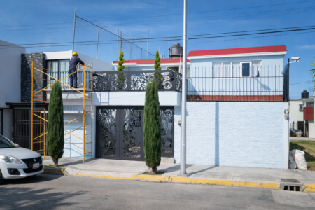 Fachada de una casa de dos niveles en remodelación con un trabajador sobre un andamio pintando o resanando la pared exterior.