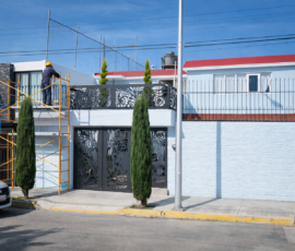 Fachada de una casa de dos niveles en remodelación con un trabajador sobre un andamio pintando o resanando la pared exterior.